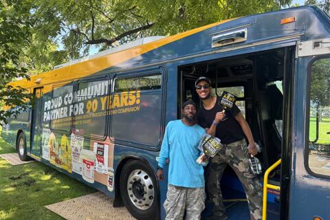 Two people standing by a community outreach bus at an outdoor event.