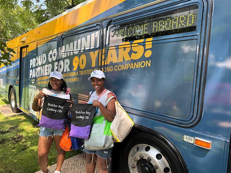 Two attendees holding giveaway bags in front of a community outreach bus.